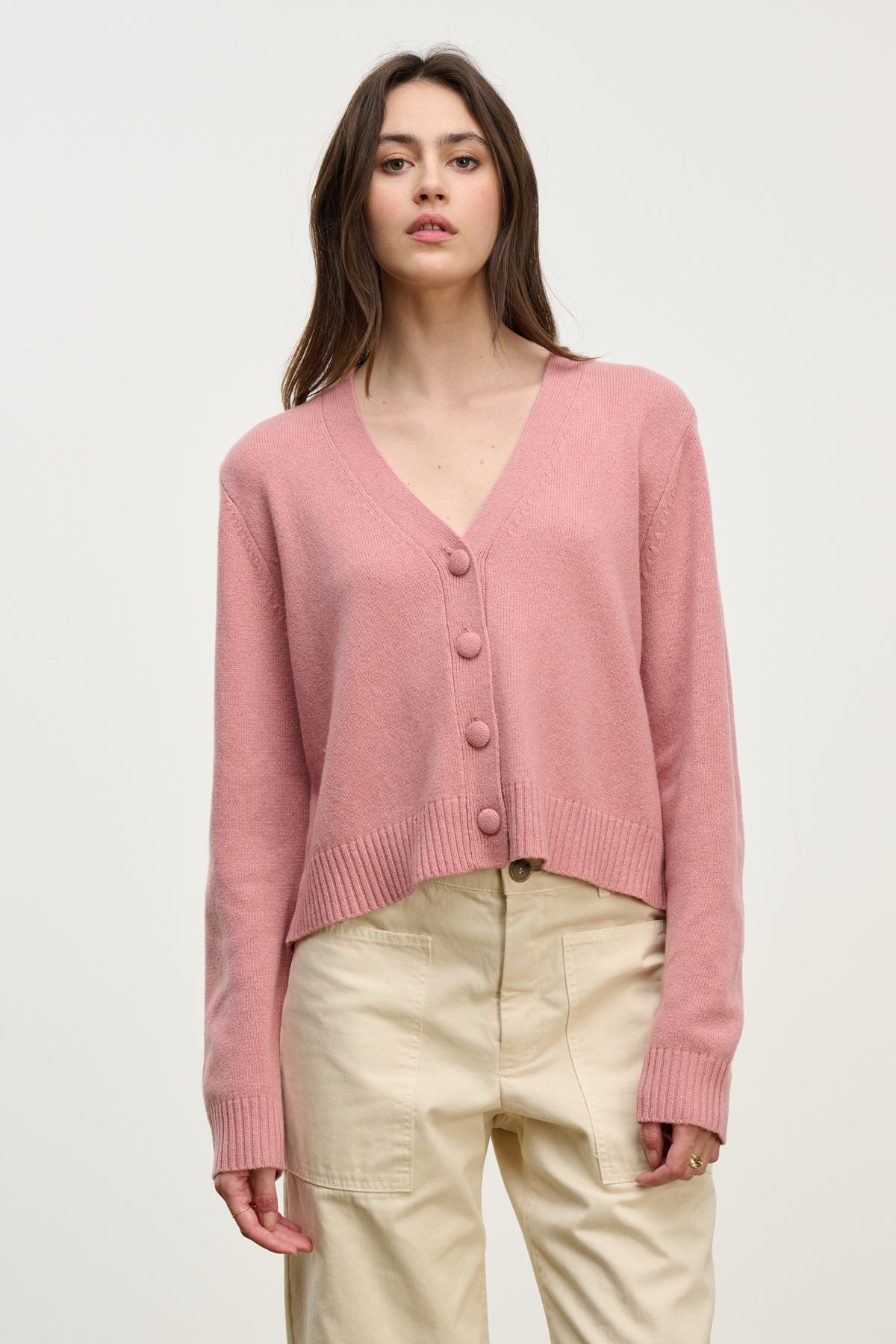 A woman wears the ETNA CARDIGAN by Velvet by Graham & Spencer, a light pink wool blend with a V-neckline, paired with high-waisted beige pants against a plain, light background.-40227054747841