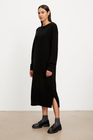 A woman stands side-profile in a plain, long black KADEN SWEATER DRESS with side split hems and black loafers, against a neutral background by Velvet by Graham & Spencer.