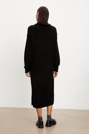 A woman from behind, wearing a KADEN SWEATER DRESS by Velvet by Graham & Spencer with a side split hem and black boots, standing against a plain white background.