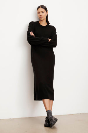 A woman in a black long-sleeve KADEN SWEATER DRESS by Velvet by Graham & Spencer with a crew neckline and boots, standing against a white background with arms crossed.