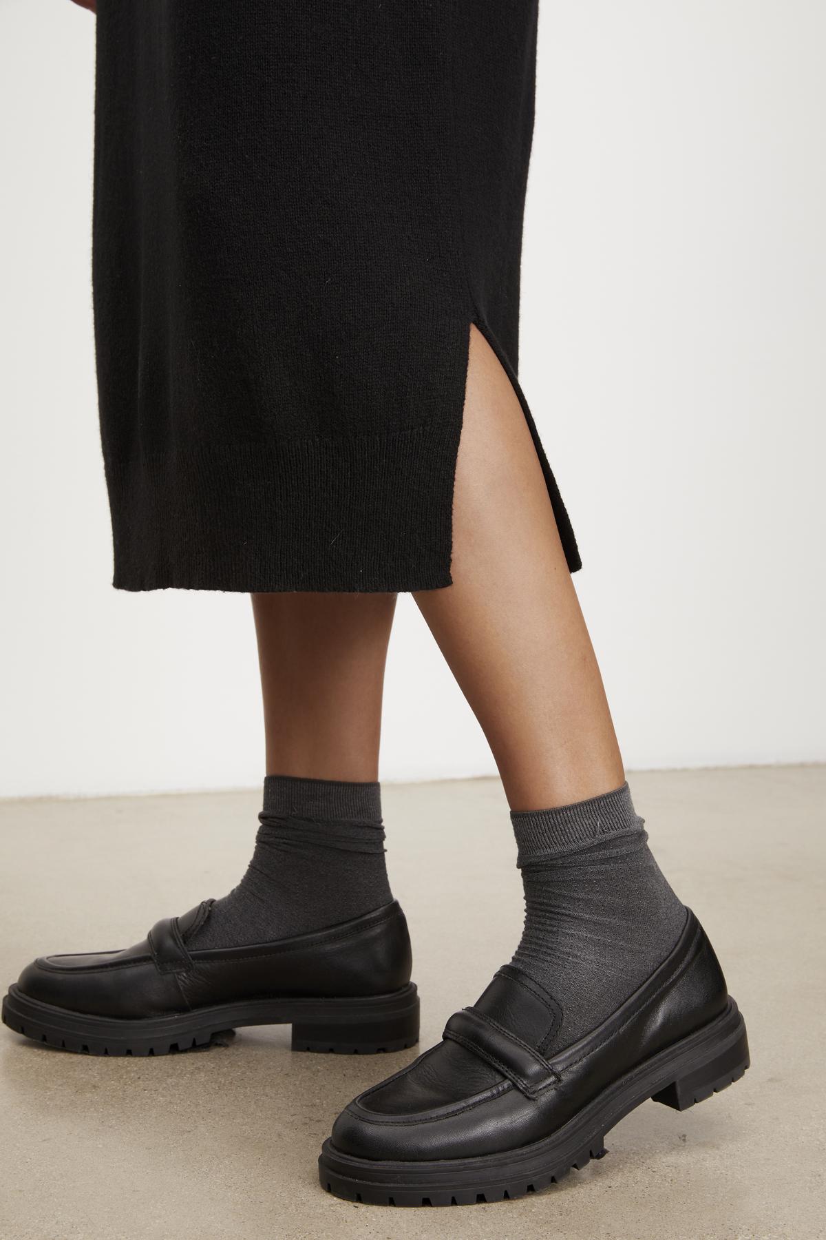 Person wearing black loafers and gray socks, standing on a light-colored floor, showcasing the versatile KADEN SWEATER DRESS by Velvet by Graham & Spencer with its stylish side slit.-37618712936641