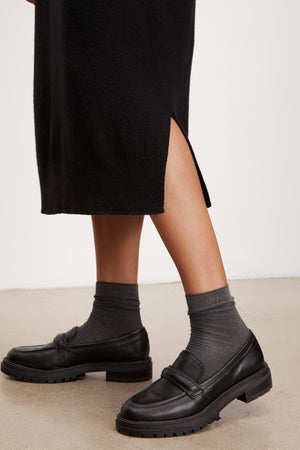 Person wearing black loafers and gray socks, standing on a light-colored floor, showcasing the versatile KADEN SWEATER DRESS by Velvet by Graham & Spencer with its stylish side slit.