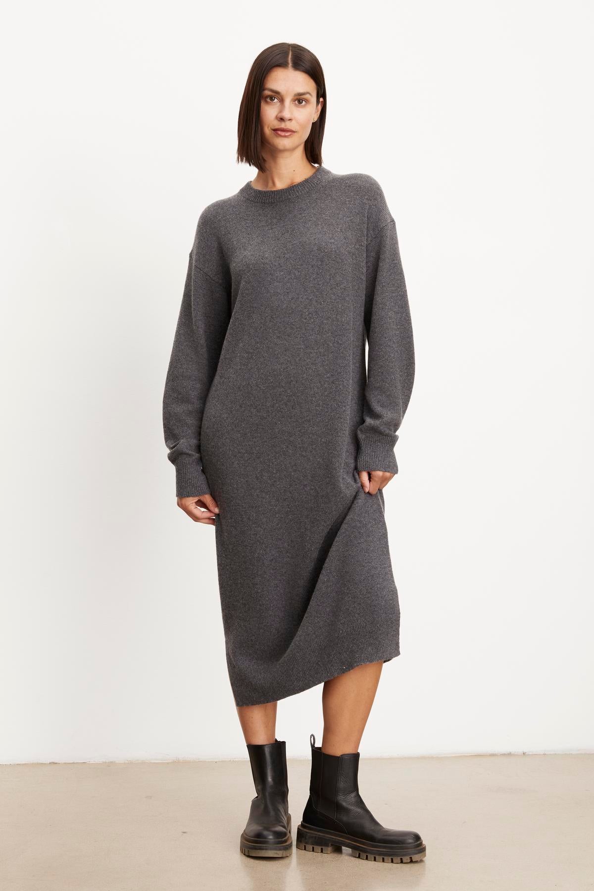   A woman standing in a minimalist setting, wearing a long, gray KADEN SWEATER DRESS with a side split hem and black combat boots from Velvet by Graham & Spencer. 