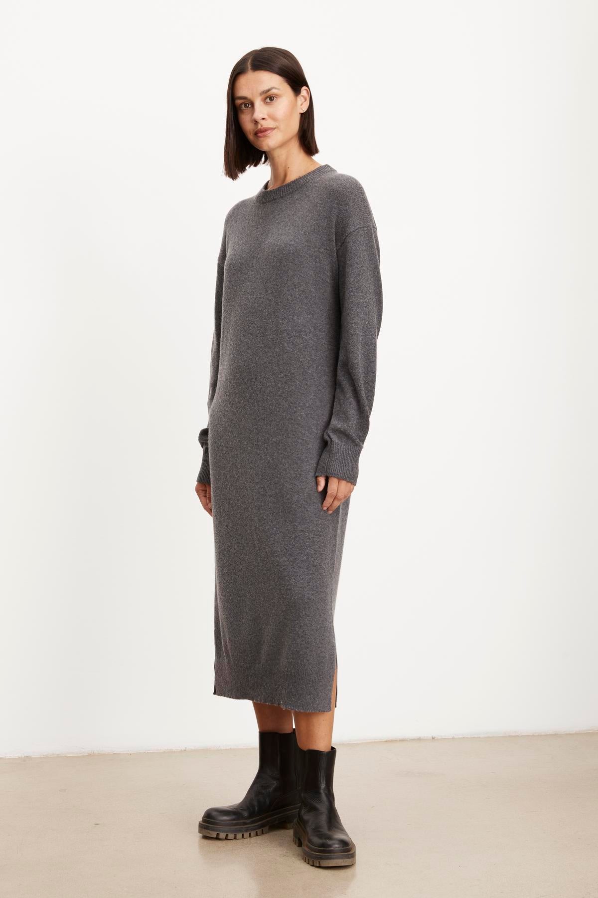   A woman stands wearing a long gray KADEN SWEATER DRESS by Velvet by Graham & Spencer with a side split hem and black combat boots, posing with her hands slightly tucked into the dress sides, against a plain white background. 