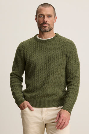 A man wears the ALONZO SWEATER by Velvet by Graham & Spencer in olive green with light beige pants, standing against a plain, light background.