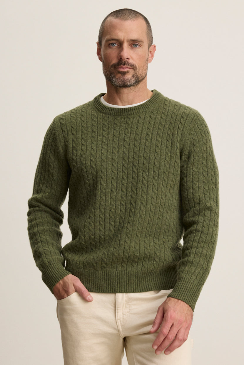 A man wears the ALONZO SWEATER by Velvet by Graham & Spencer in olive green with light beige pants, standing against a plain, light background.