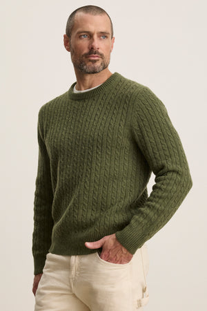 A man with short hair and a beard wears the olive green ALONZO SWEATER by Velvet by Graham & Spencer and light beige pants, standing against a plain background.