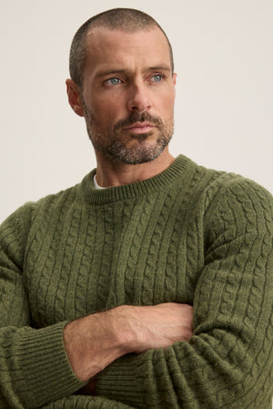 A man with short hair and a beard wears the green ALONZO SWEATER by Velvet by Graham & Spencer, standing with arms crossed and gazing slightly to the side.