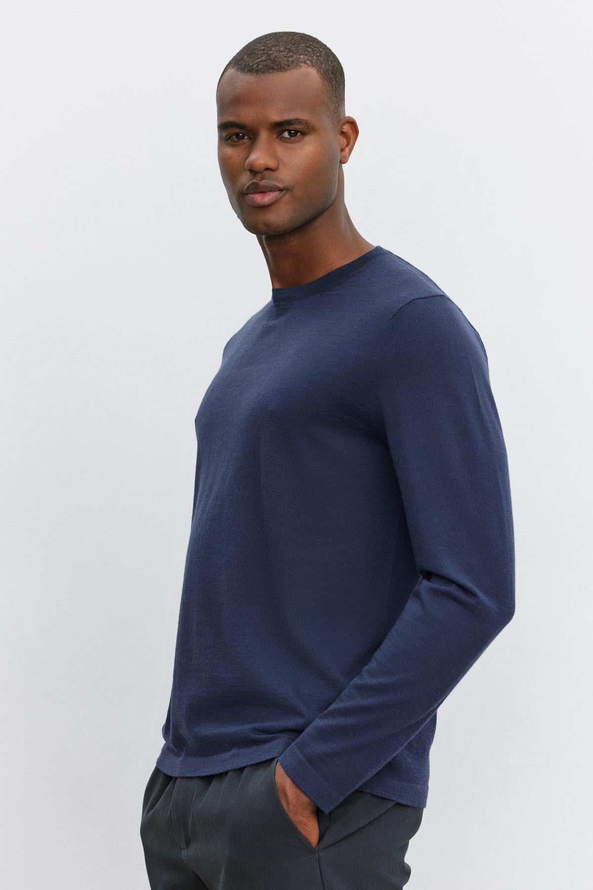   A man wearing the ALVIN SWEATER, a long-sleeve navy blue top from Velvet by Graham & Spencer, and dark pants, stands with his hand in his pocket against a plain light background. 