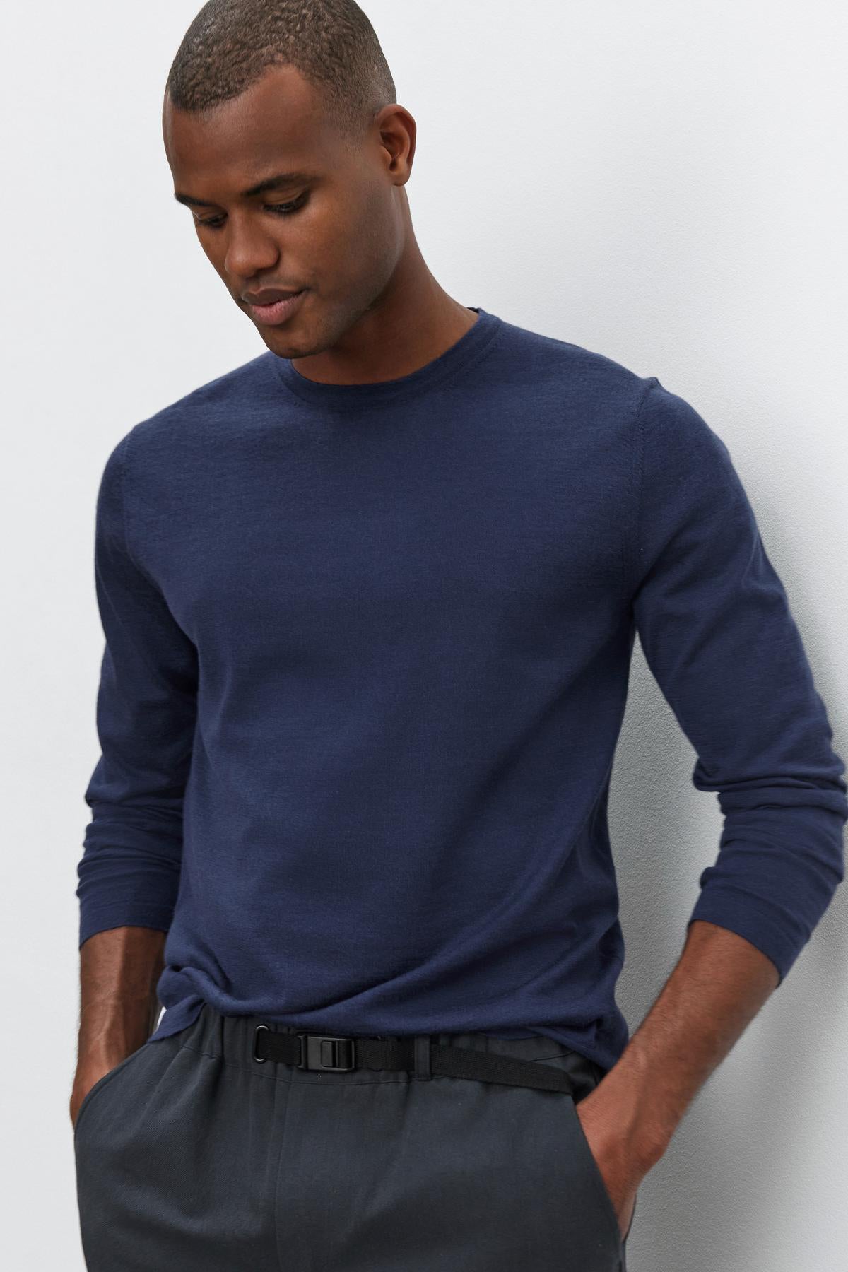   A man wearing an ALVIN SWEATER, a navy blue long-sleeve shirt by Velvet by Graham & Spencer, and dark gray pants stands against a plain light-colored background, looking downwards with his hands in his pockets. 