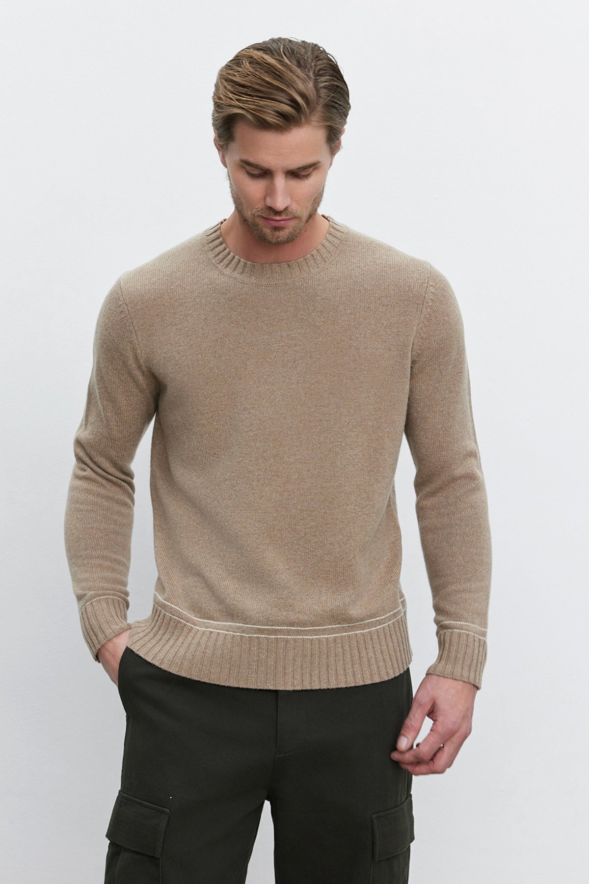   A man wearing the ATTICUS SWEATER, a beige wool blend crew neck sweater by Velvet by Graham & Spencer, and dark pants stands against a plain background with his hands in his pockets. 