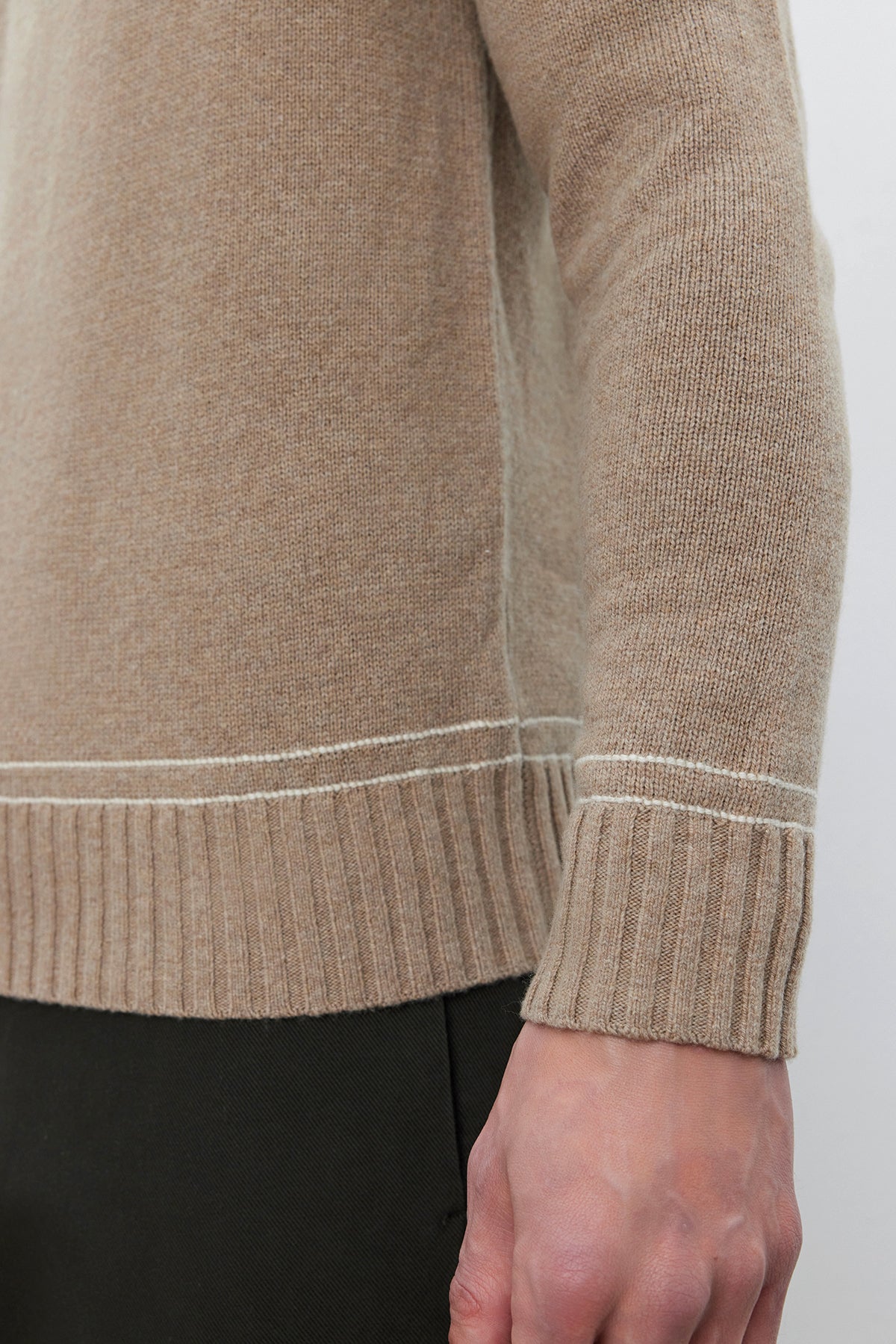   A close-up of a person's arm wearing the ATTICUS SWEATER, a beige knit crew neck made from a wool blend with ribbed cuffs and hem, by Velvet by Graham & Spencer. The person is standing against a neutral background. 