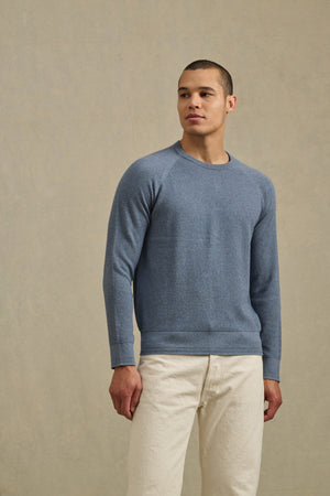 A man stands against a plain beige background, wearing the AUGIE SWEATER by Velvet by Graham & Spencer—a blue cotton-cashmere crew neck—paired with off-white pants.