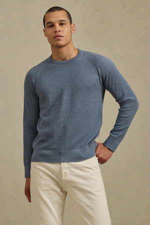 A man stands against a neutral background, wearing the AUGIE SWEATER by Velvet by Graham & Spencer—a textured blue cotton-cashmere crewneck—paired with off-white pants, one hand resting on his hip.