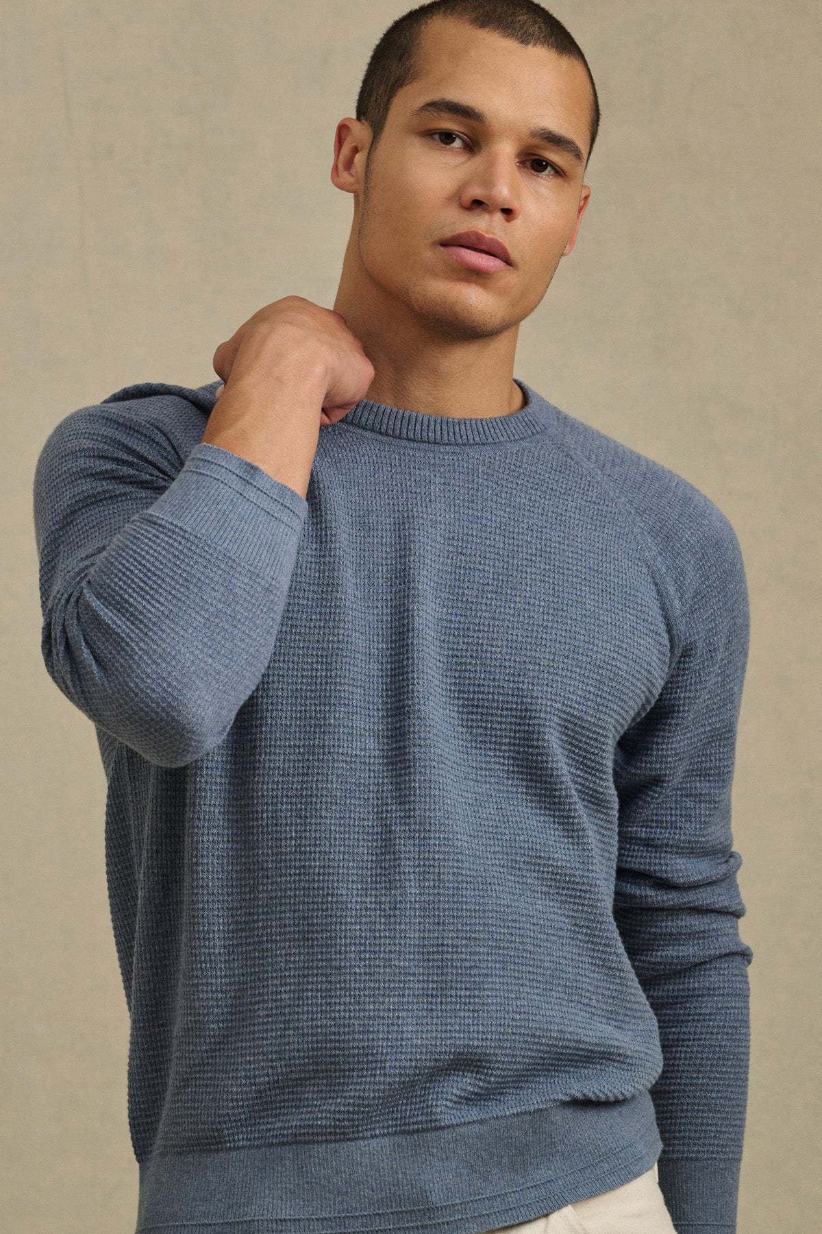 A man stands against a beige background, wearing Velvet by Graham & Spencer's AUGIE SWEATER—a textured blue cotton-cashmere crewneck—paired with light pants, with one hand resting on his shoulder.-40850648203457