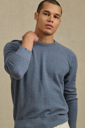 A man stands against a beige background, wearing Velvet by Graham & Spencer's AUGIE SWEATER—a textured blue cotton-cashmere crewneck—paired with light pants, with one hand resting on his shoulder.