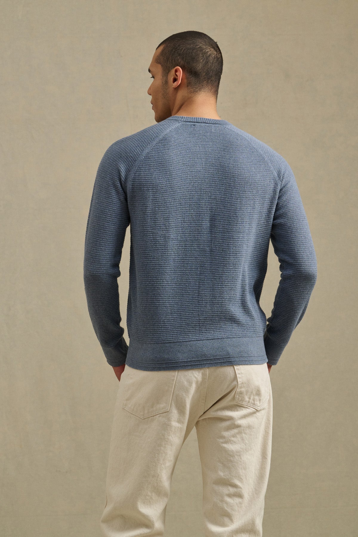 A man faces away from the camera, wearing the Velvet by Graham & Spencer AUGIE SWEATER—a textured blue cotton-cashmere crewneck—paired with cream pants against a neutral background.-40850648105153