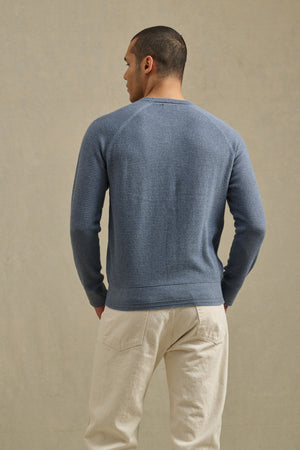 A man faces away from the camera, wearing the Velvet by Graham & Spencer AUGIE SWEATER—a textured blue cotton-cashmere crewneck—paired with cream pants against a neutral background.