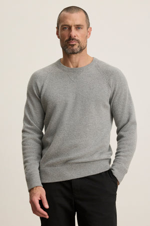A man with short hair and a beard wears the CORTEZ SWEATER by Velvet by Graham & Spencer in light gray, paired with black pants, standing against a plain light background.
