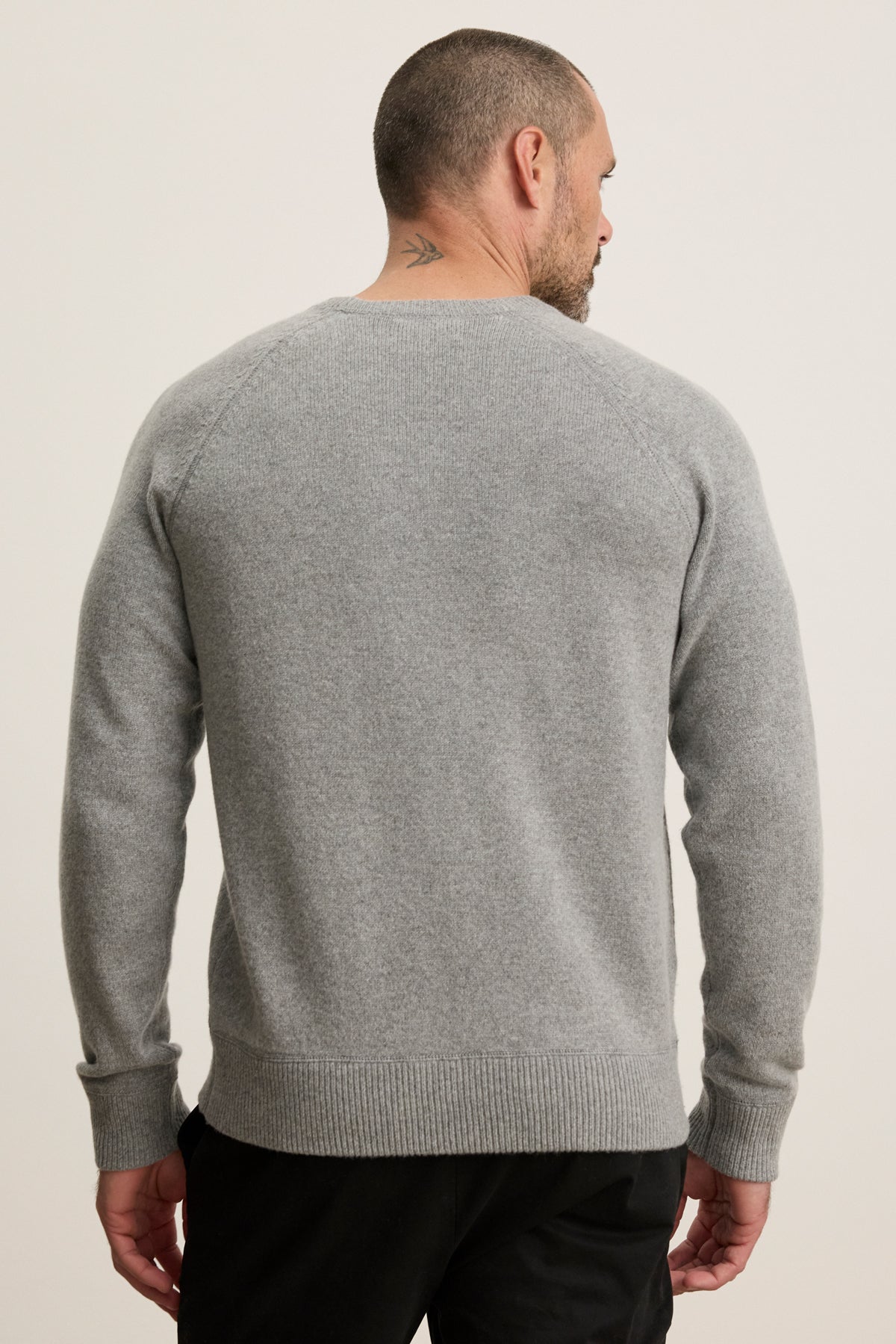 A man with a short haircut and a small neck tattoo is wearing the Velvet by Graham & Spencer CORTEZ SWEATER in light grey and black pants, facing away from the camera.-40054234546369