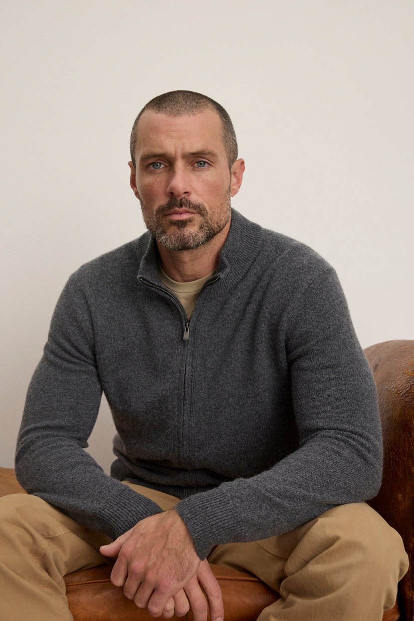 A man with short hair and a beard sits on a brown chair against a plain light background, wearing beige pants and the Velvet by Graham & Spencer DALLEN FULL ZIP SWEATER, crafted from a soft wool blend for extra comfort.