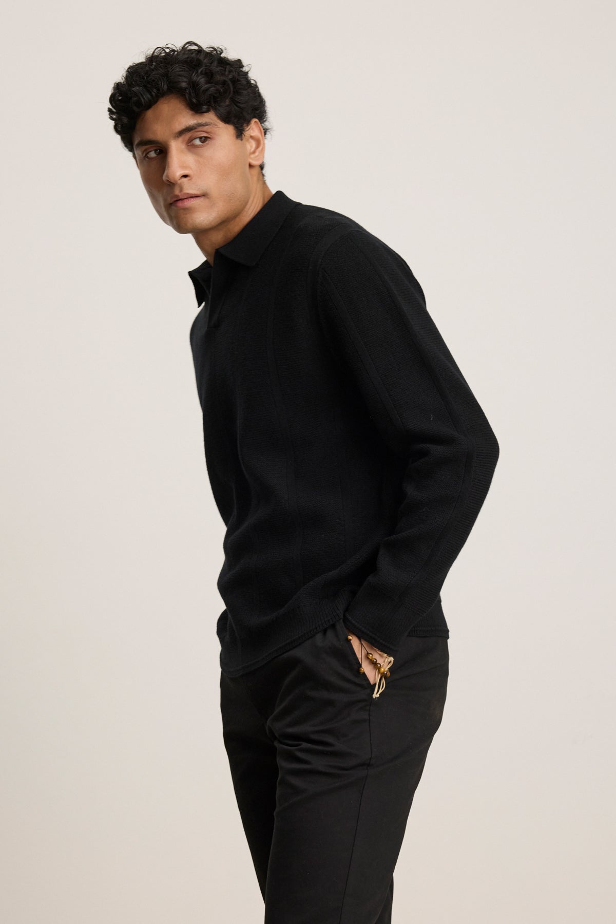 A man with curly black hair wears the Velvet by Graham & Spencer REDDING SWEATER POLO and black pants against a plain light background—a perfect look for versatile layering.
