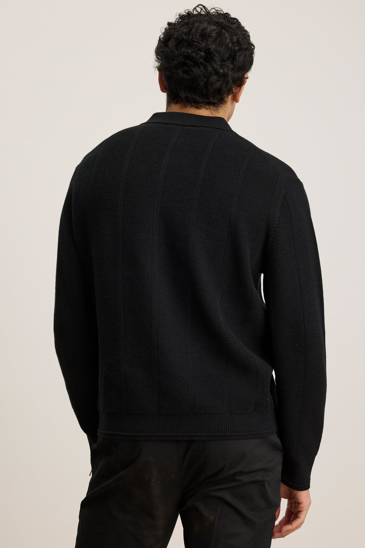 A man with dark curly hair is seen from behind, wearing the REDDING SWEATER POLO by Velvet by Graham & Spencer and black pants, standing against a plain light background.-40054247915713