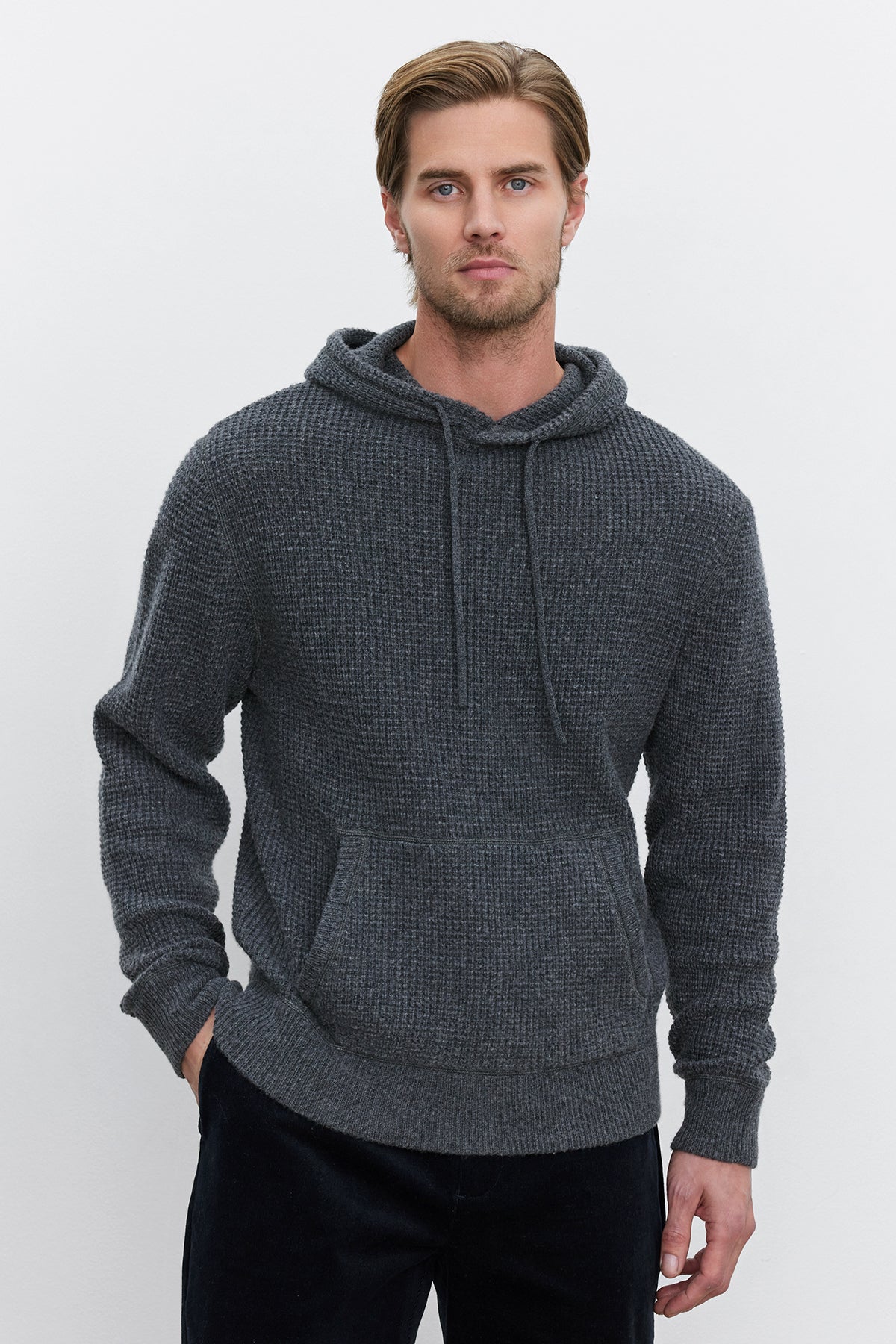   A man with light brown hair and a beard wearing the SHANE SWEATER HOODIE by Velvet by Graham & Spencer and black pants stands against a plain white background. 