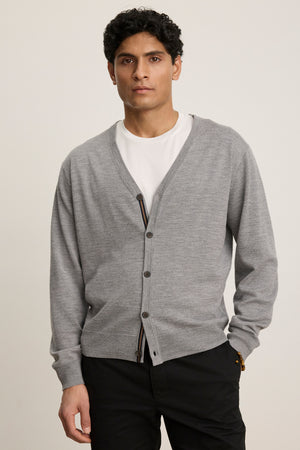 A man wears the SILVIO CARDIGAN by Velvet by Graham & Spencer over a white t-shirt and black pants, standing against a plain background.