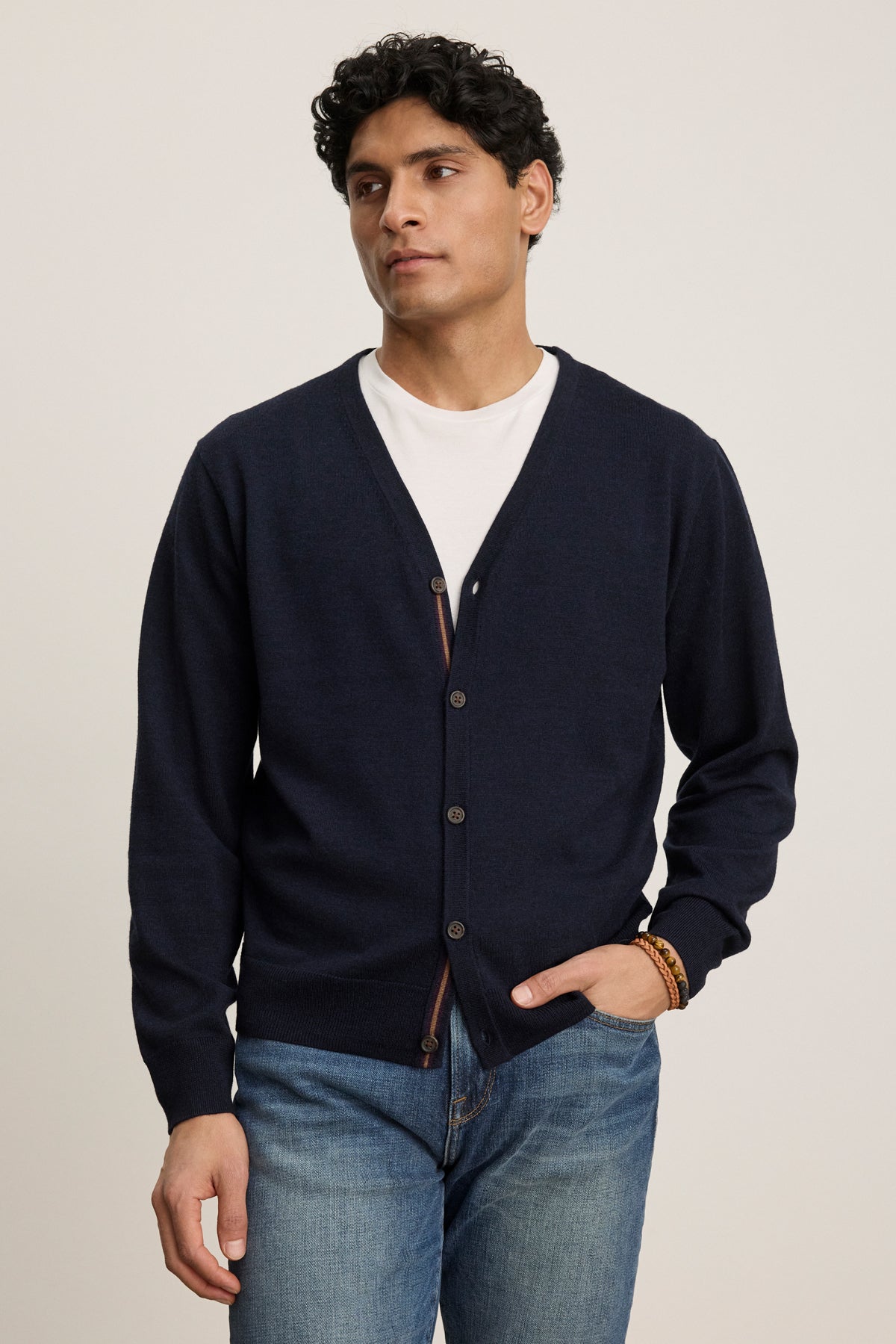 A man wearing the SILVIO CARDIGAN by Velvet by Graham & Spencer over a white t-shirt and blue jeans stands with one hand in his pocket.-39543530717377