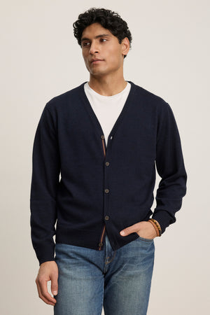 A man wearing the SILVIO CARDIGAN by Velvet by Graham & Spencer over a white t-shirt and blue jeans stands with one hand in his pocket.