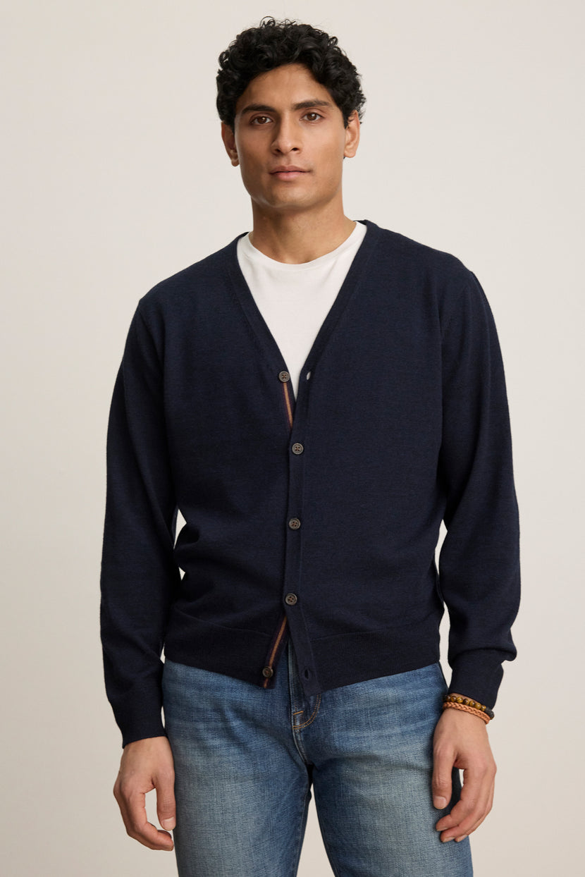 A person wears the SILVIO CARDIGAN by Velvet by Graham & Spencer—a navy blue premium wool cardigan with ribbed trims—over a white T-shirt and blue jeans, standing against a plain light background.
