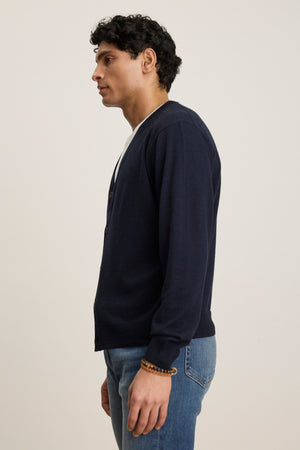 A man stands in profile against a plain background, wearing the Velvet by Graham & Spencer SILVIO CARDIGAN in navy with ribbed trims over a white shirt and blue jeans.