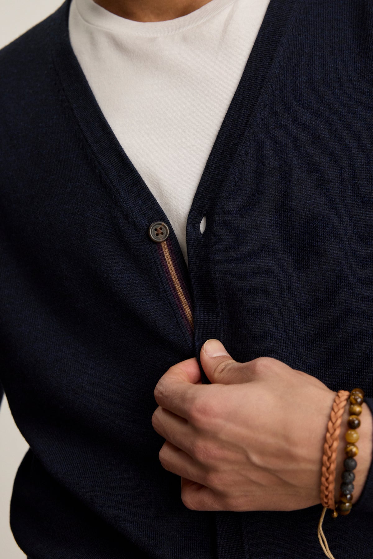 Wearing the SILVIO CARDIGAN by Velvet by Graham & Spencer, a person in a white t-shirt fastens a button, accessorized with a brown braided bracelet and beaded wristbands.-39543530586305