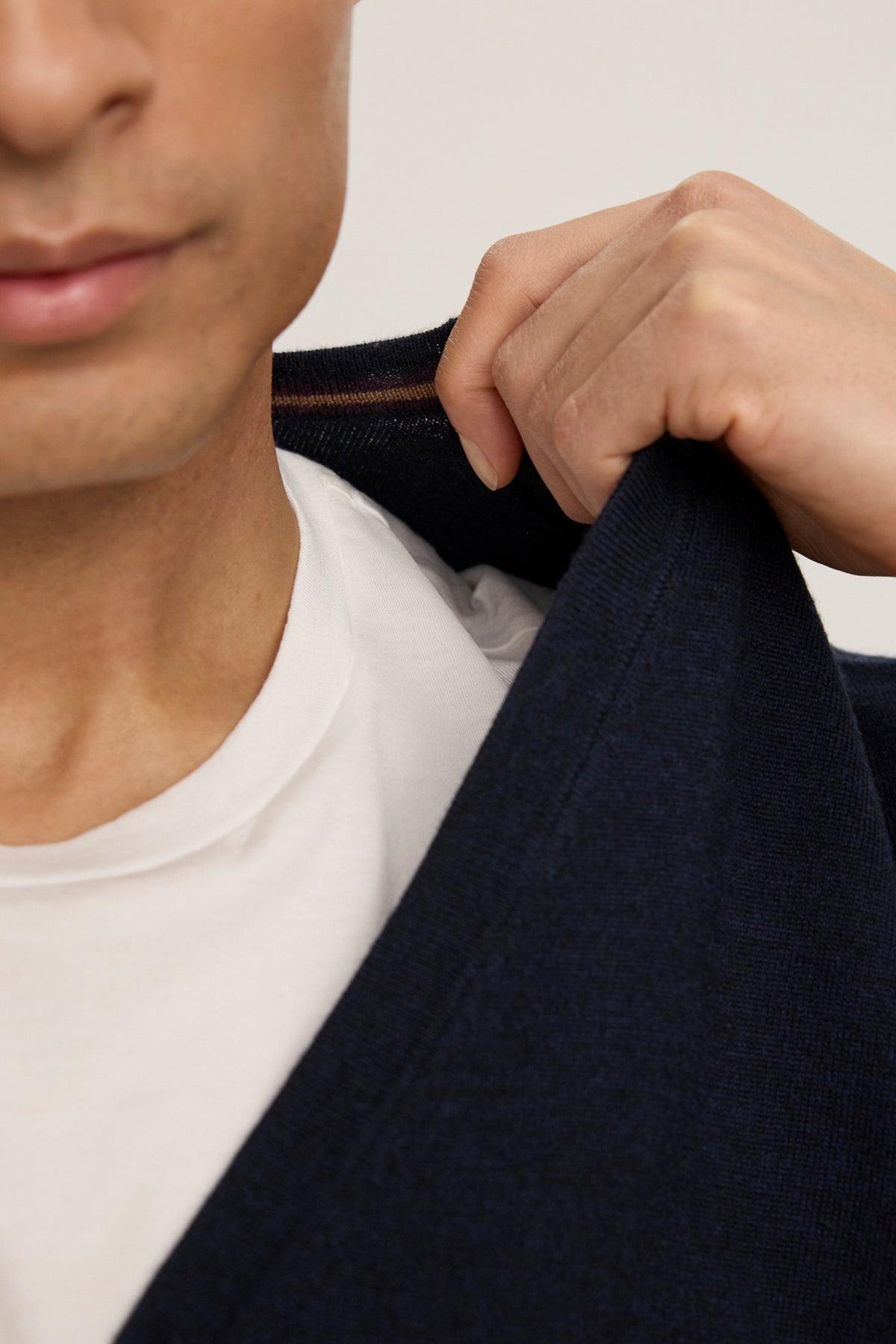 A person wearing a white t-shirt and the SILVIO CARDIGAN by Velvet by Graham & Spencer, a dark blue V-neck cardigan with ribbed trims, holding the sweater open at the collar with one hand.-39543530520769