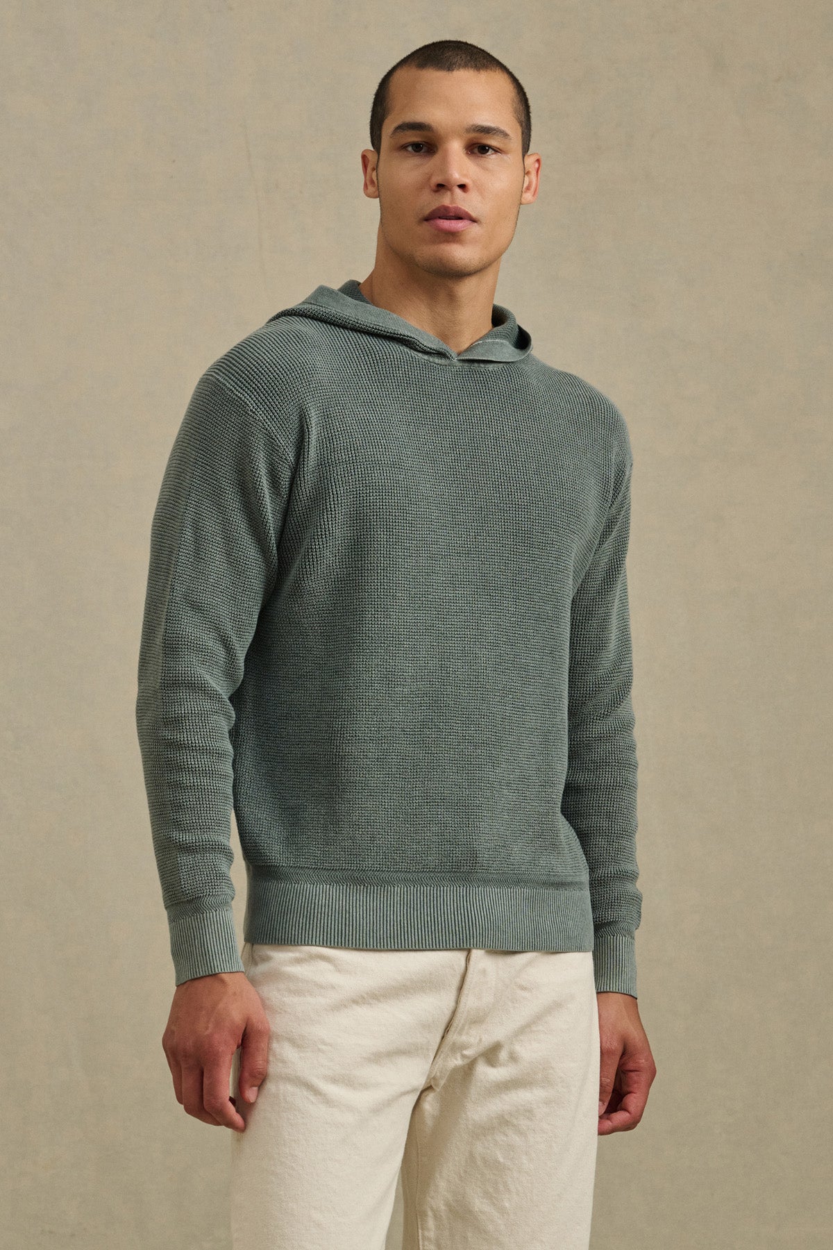 A man stands facing forward in a Velvet by Graham & Spencer WILFRED SWEATER HOODIE and off-white pants, set against a neutral background.