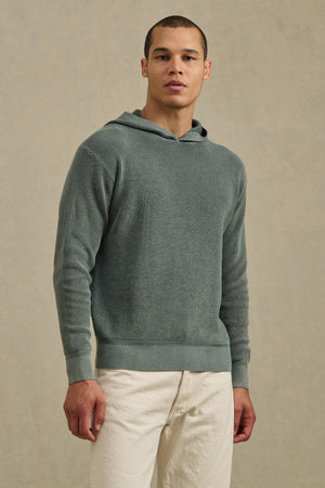 A man stands facing forward in a Velvet by Graham & Spencer WILFRED SWEATER HOODIE and off-white pants, set against a neutral background.