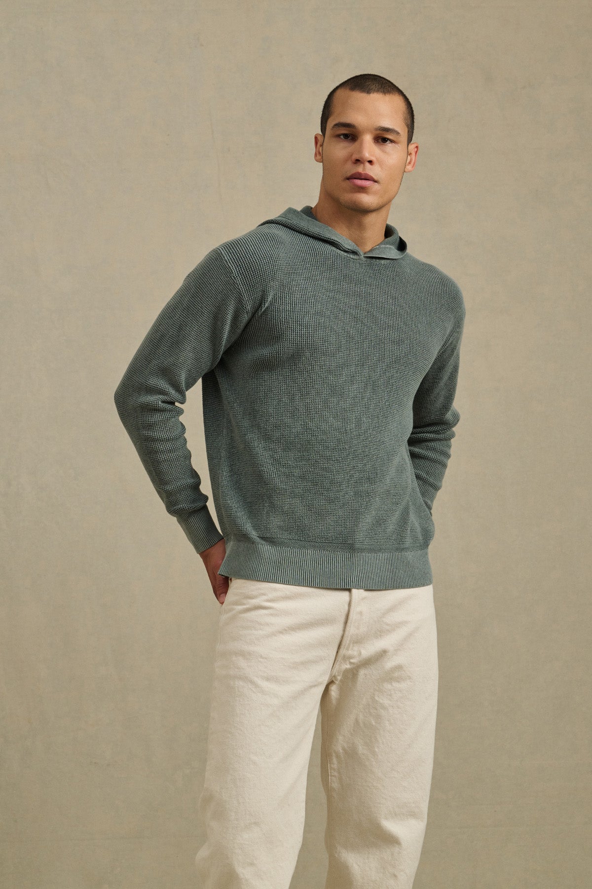 A man stands against a plain beige background wearing the WILFRED SWEATER HOODIE by Velvet by Graham & Spencer and off-white pants, with his hands behind his back.-41009083384001