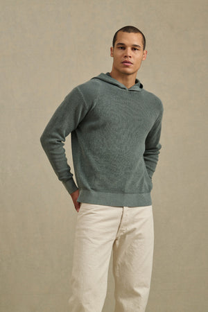 A man stands against a plain beige background wearing the WILFRED SWEATER HOODIE by Velvet by Graham & Spencer and off-white pants, with his hands behind his back.