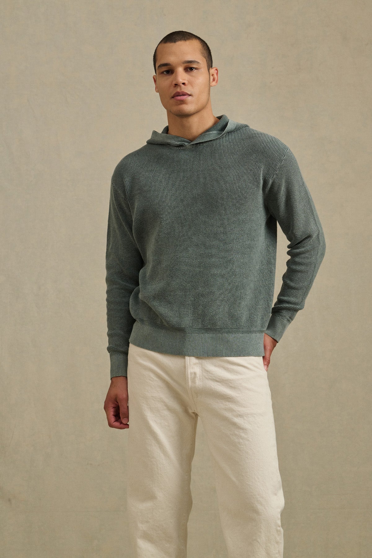 A man stands facing forward in a muted green WILFRED SWEATER HOODIE by Velvet by Graham & Spencer and off-white pants, set against a plain, light-colored background.