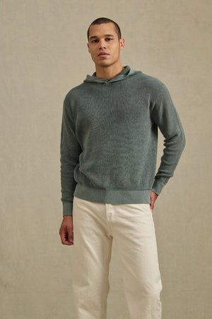 A man stands facing forward in a muted green WILFRED SWEATER HOODIE by Velvet by Graham & Spencer and off-white pants, set against a plain, light-colored background.
