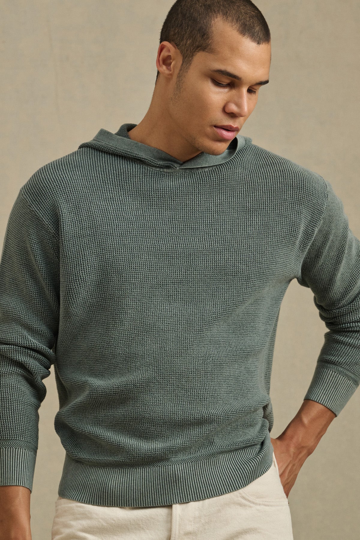 Wearing the WILFRED SWEATER HOODIE by Velvet by Graham & Spencer in textured sage green cotton knit and light beige pants, a person stands against a plain background looking down.