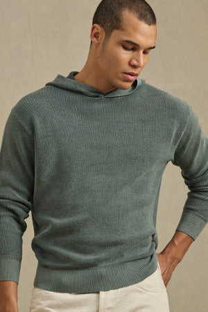 Wearing the WILFRED SWEATER HOODIE by Velvet by Graham & Spencer in textured sage green cotton knit and light beige pants, a person stands against a plain background looking down.