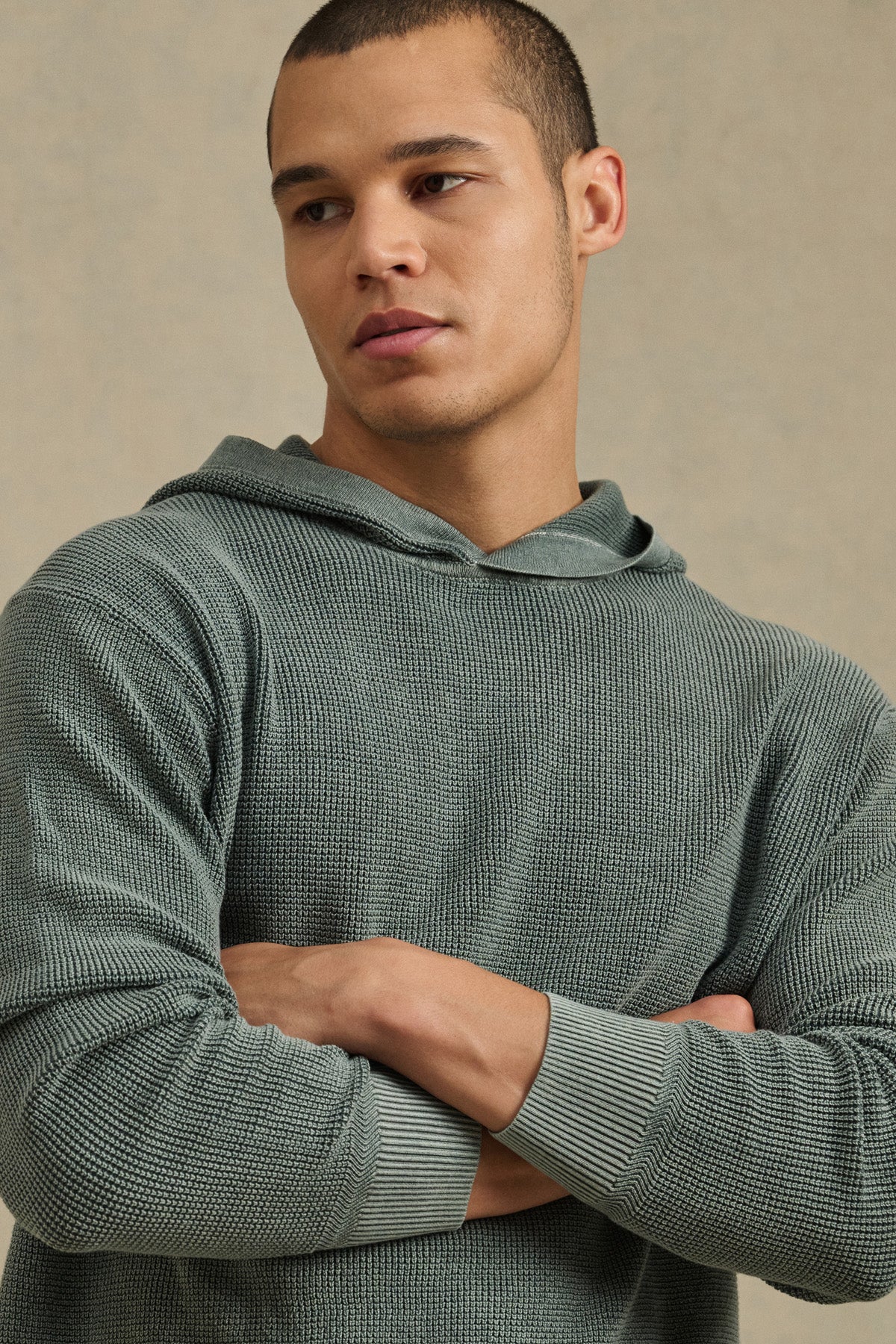 A man with short hair wears the WILFRED SWEATER HOODIE by Velvet by Graham & Spencer in textured cotton knit light green, standing with arms crossed and looking to the side.