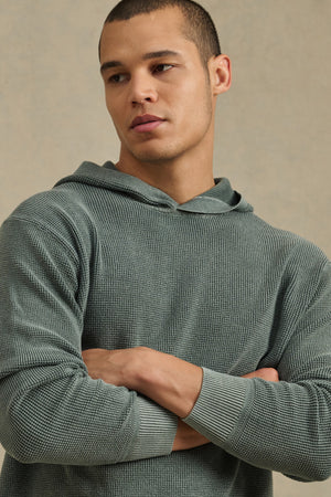 A man with short hair wears the WILFRED SWEATER HOODIE by Velvet by Graham & Spencer in textured cotton knit light green, standing with arms crossed and looking to the side.