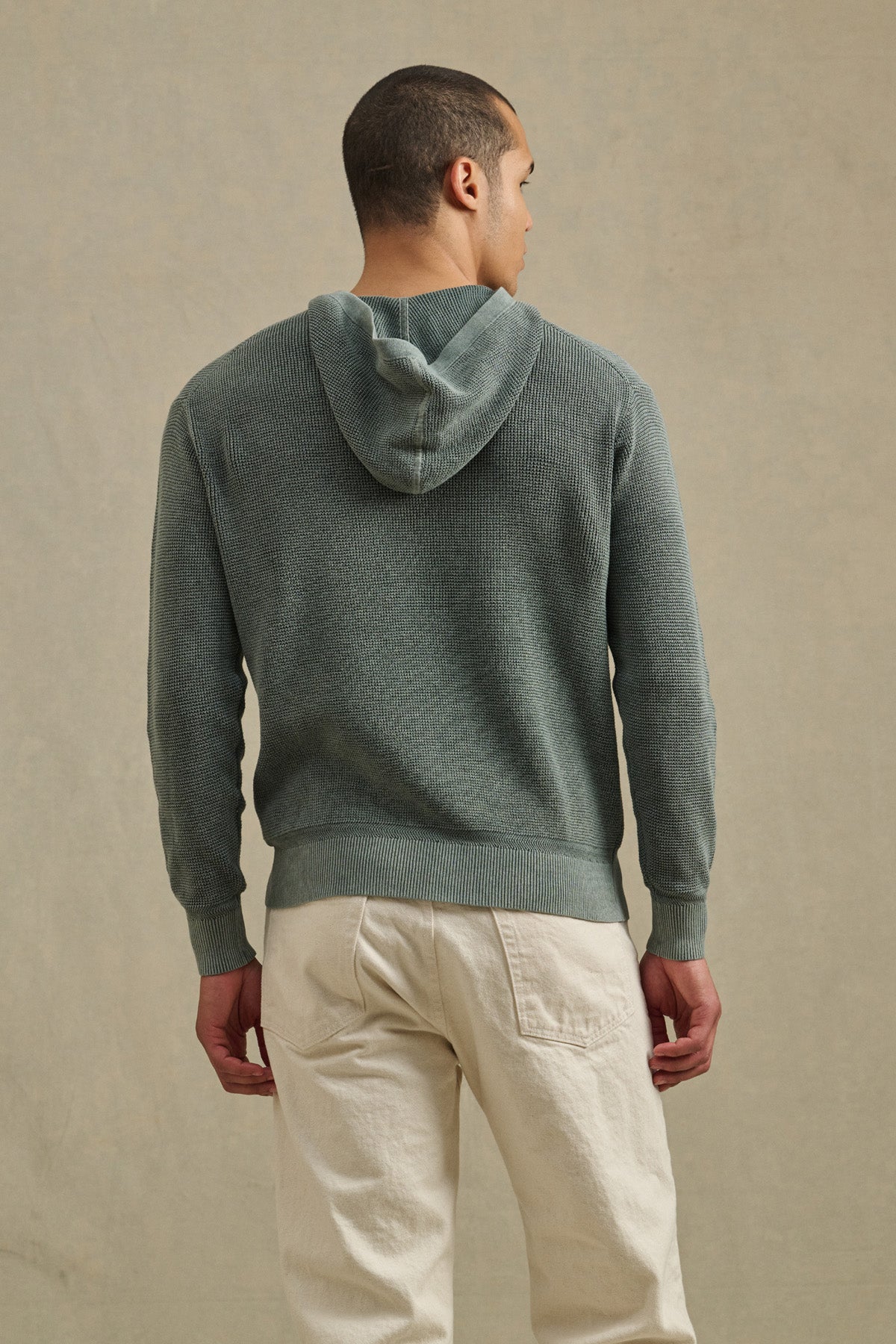 A man faces away, wearing the WILFRED SWEATER HOODIE by Velvet by Graham & Spencer—a textured green cotton knit hoodie—paired with light pants, perfect loungewear against a plain, neutral backdrop.