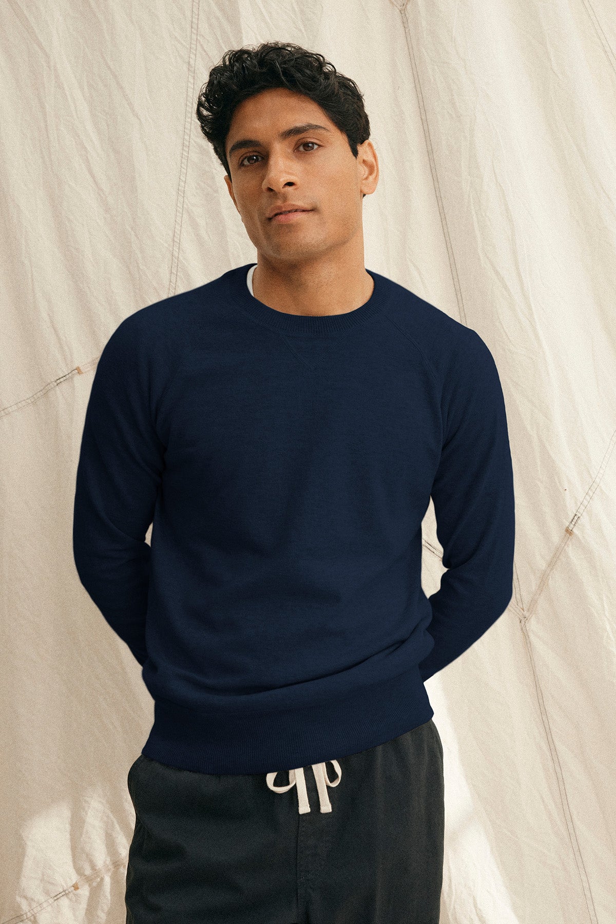 A man stands before a light fabric backdrop wearing the DARIO SWEATER by Velvet by Graham & Spencer, a navy blue cotton cashmere crewneck, paired with black drawstring pants.