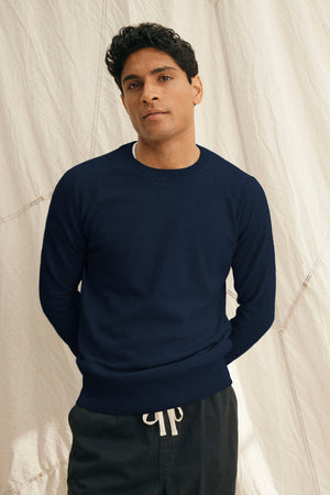 A man stands before a light fabric backdrop wearing the DARIO SWEATER by Velvet by Graham & Spencer, a navy blue cotton cashmere crewneck, paired with black drawstring pants.