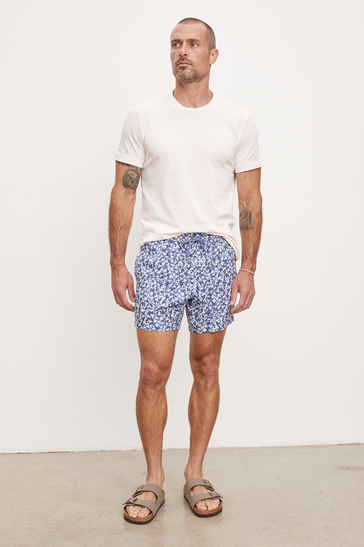   A man standing against a white background wearing a white t-shirt, Velvet by Graham & Spencer RICARDO SWIM SHORTS, and brown sandals. He has tattoos on his arms and is looking to the side. 