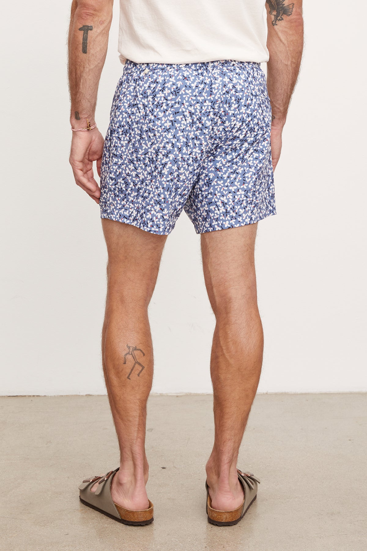 Man in Velvet by Graham & Spencer's RICARDO SWIM SHORT and brown flip-flops, standing with his back to the camera, displaying a tattoo on his right calf.-36918541910209